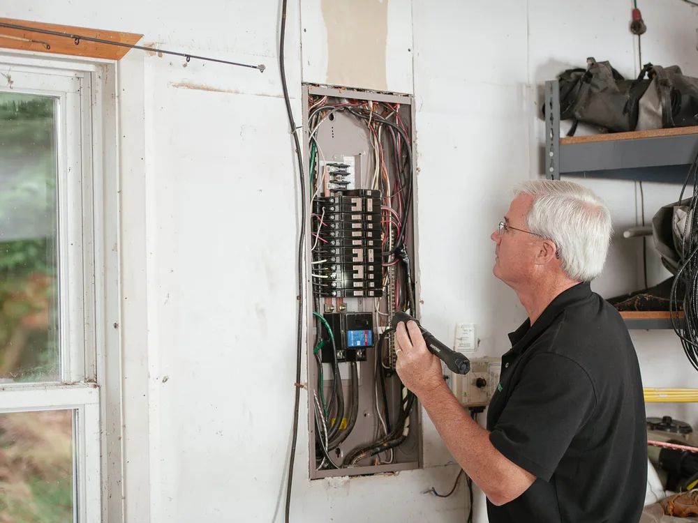 Electrician inspecting an open residential breaker panel with a flashlight during a panel upgrade evaluation